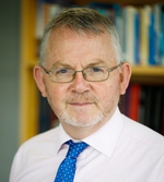 A middle-aged man with grey hair, a beard, and glasses, wearing a light pink shirt and blue polka-dot tie, stands in front of a bookcase filled with books—one of the Global Cancer Update Programme Experts.