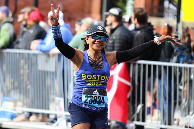 A smiling marathon runner in a Boston 2022 vest, sunglasses, and cap raises her arms in celebration, flashing a peace sign as she nears the finish line, with spectators behind a metal barrier.