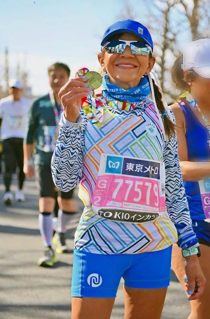 A smiling marathon runner wearing sunglasses, a cap, and a colourful outfit holds up a finisher's medal. Other runners and trees are visible in the background. The runner's bib reads 77579 and 東京メトロ (Tokyo Metro).