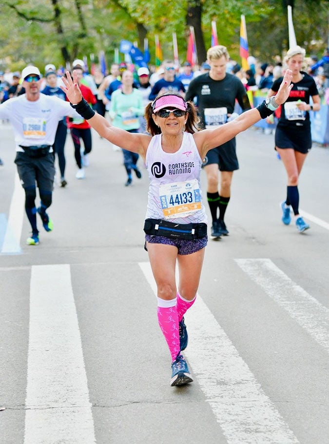 A woman with her arms raised runs in a marathon, wearing a white vest, pink visor, and pink knee-high socks. Other runners are visible behind her on a road lined with trees and flags.