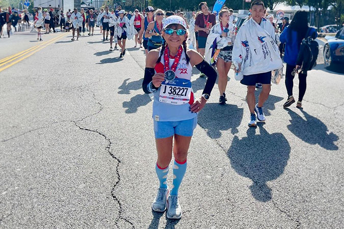 A smiling marathon runner in blue kit stands on a sunlit road holding a medal, with other participants and spectators walking behind her. She wears sunglasses, a headband, and race number J38227.