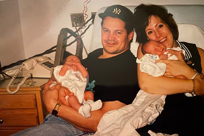 A man and woman sit closely together on a bed, each holding a newborn baby. The man wears a dark T-shirt and a baseball cap, while the woman smiles, holding her baby gently in her arms. A phone and lamp are on the bedside table.