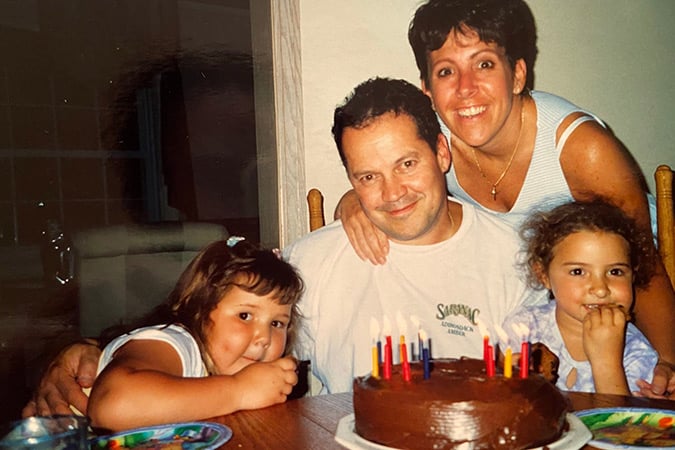 A man sits at a table with two young girls and a woman standing behind him. They are smiling at the camera with a chocolate birthday cake decorated with colourful candles in front of them.