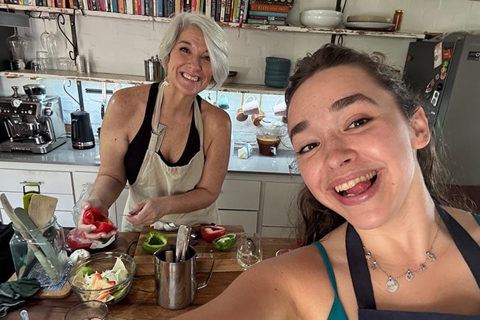 Two women smile for a selfie in a cosy kitchen while preparing vegetables. One woman chops peppers on a chopping board, and the other holds the camera, both wearing aprons and surrounded by kitchen tools and ingredients.