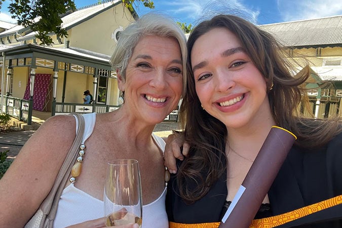 Two women smiling outdoors; one holds a drink and the other, dressed in a graduation gown, holds a rolled certificate. They stand in front of a building with a veranda and trees under a blue sky.