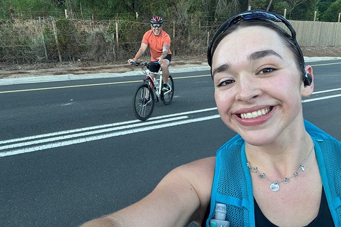 A woman in sports gear smiles for a selfie on a road, whilst a man wearing a helmet and orange shirt rides a bicycle behind her. Trees and a fence line the background.