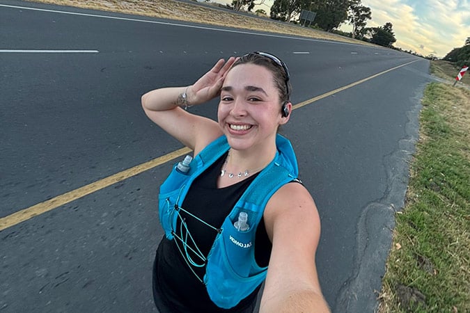 A smiling woman wearing a blue hydration vest and earphones takes a selfie whilst jogging on the side of an empty road, saluting with one hand. Trees and grass line the edge of the road under a partly cloudy sky.