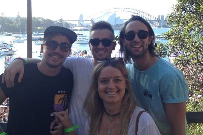 A group of three men and one woman smiling and posing together outdoors with Sydney Opera House and Harbour Bridge in the background on a sunny day.