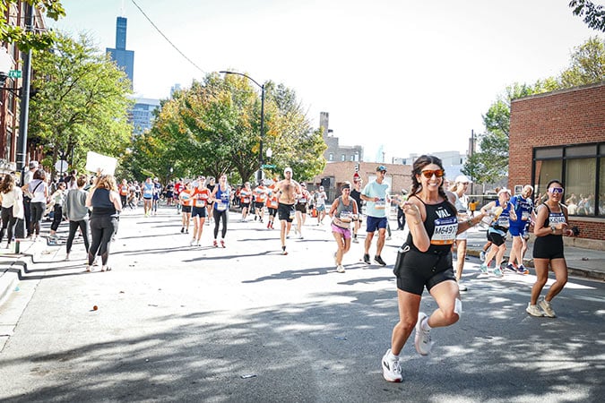 Runners participate in a marathon on a sunny city street, with spectators lining the pavements. One runner in the foreground smiles and makes a peace sign towards the camera. Trees and buildings are visible in the background.