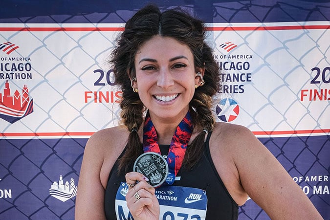 Val – A smiling woman holds up a finisher's medal in front of a Chicago Marathon backdrop, wearing a race number and sports kit.