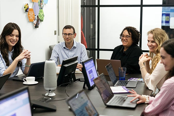 A group of five people sit around a conference table with laptops, smiling and conversing during a meeting in a modern office setting.