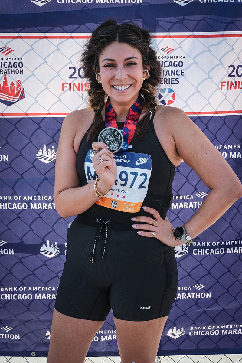 A smiling woman stands in front of a Chicago Marathon backdrop, holding a finisher’s medal. She is wearing a race bib numbered 1972, a black athletic outfit, and a red ribbon round her neck.