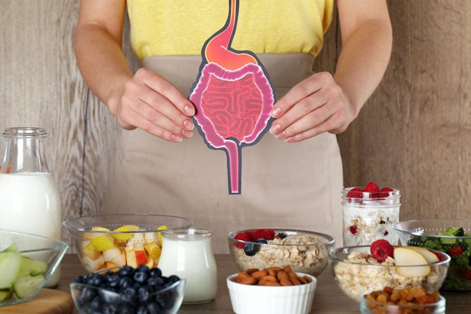 A person holding an illustrated cut-out of a digestive system stands behind a table with healthy foods, highlighting the importance of diet and sharing information about bowel colorectal cancer prevention.