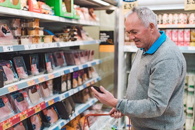 An older man with grey hair stands in a supermarket aisle, holding a shopping basket and looking at his mobile phone for information about bowel colorectal cancer while shopping for packaged meats on the refrigerated shelves.