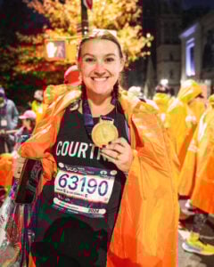 A smiling woman wearing a race number and medal stands outside at night, draped in an orange poncho among other runners in similar attire, celebrating after finishing a marathon.