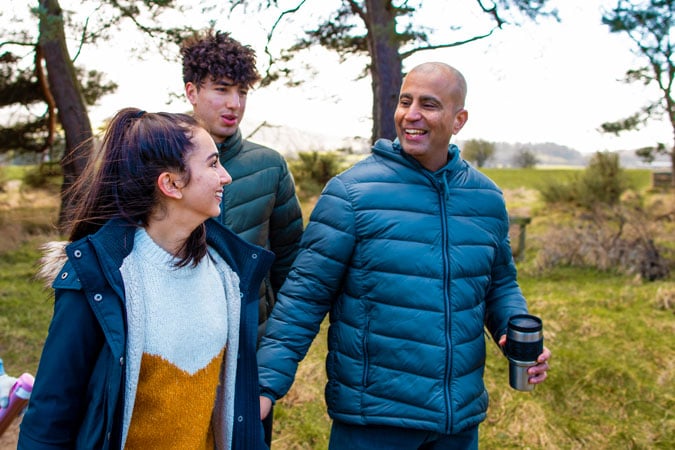 Three people wearing jackets smile and walk together on a nature trail, chatting happily. The man in front holds a travel mug as they enjoy the fresh air and share information about bowel colorectal cancer along the way. Trees and grassy fields surround them.