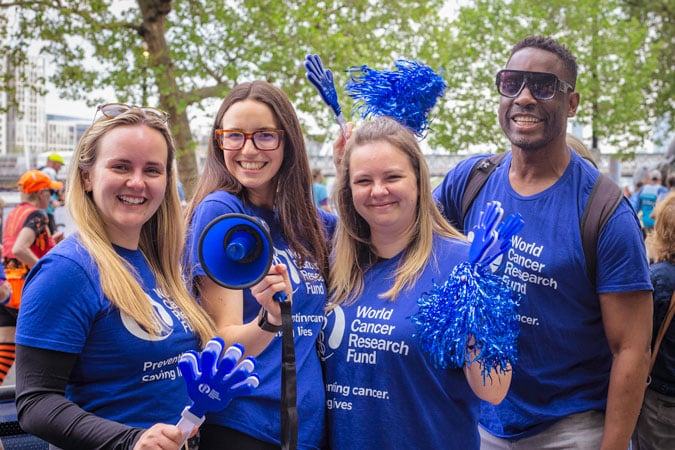 Four smiling people wearing blue World Cancer Research Fund shirts pose outdoors, holding blue pom-poms, plastic hands, and a megaphone, with trees and a bright atmosphere in the background.