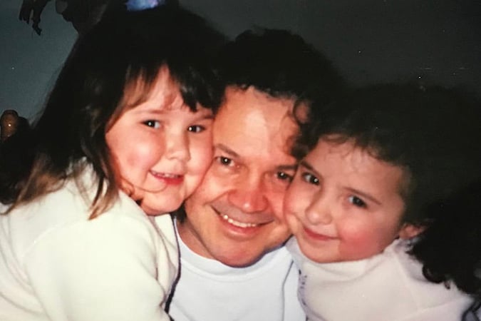 A man smiles as he hugs two young girls, one on each side, all close together and looking at the camera. The girls appear happy and affectionate, wearing light-coloured clothing.