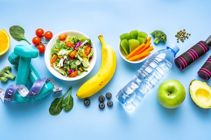 A flat lay of healthy lifestyle items: dumbbells with a measuring tape, a bowl of salad, fresh fruit and vegetables, a bottle of water, and a skipping rope on a blue background.