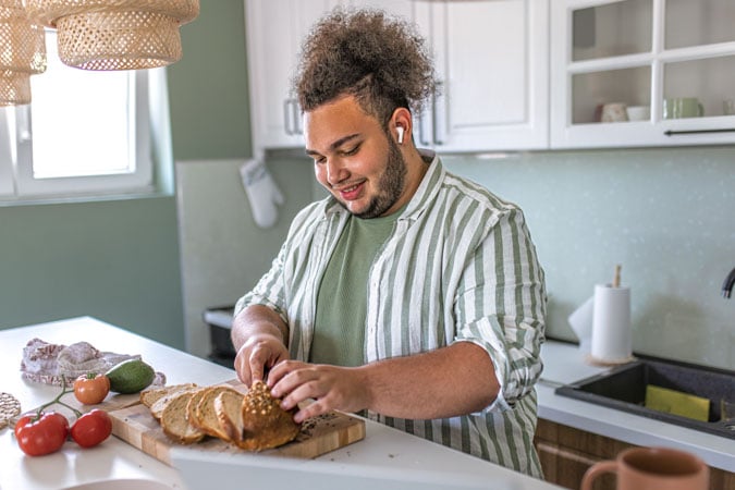 A person wearing a striped shirt and wireless earphones slices bread on a chopping board in a bright kitchen, surrounded by vegetables and kitchenware, while listening to information about bowel colorectal cancer.