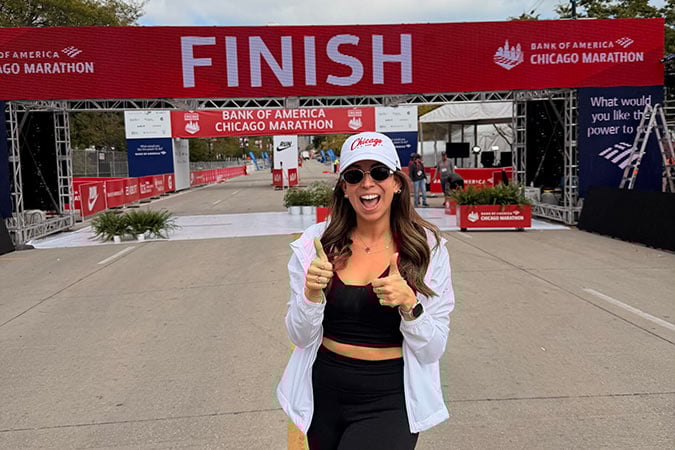 A smiling woman in sportswear and a white cap stands near the finish line of the Bank of America Chicago Marathon, giving two thumbs up. The red finish banner and race signage are visible in the background.