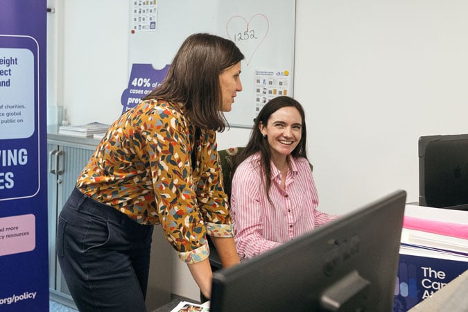 Two women in an office, one standing and one sitting at a desk with a computer. They are smiling and talking. Posters and charts are visible in the background.