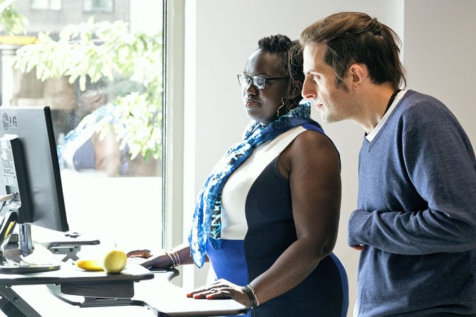 Two people stand at a desk looking intently at a computer monitor. Sunlight streams through a window nearby, and there is a banana and an apple on the desk.
