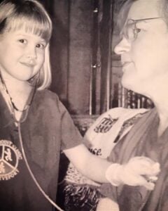 A young girl wearing a stethoscope plays doctor with a woman who is smiling and wearing glasses. The girl holds the stethoscope to the woman's chest. The photo is black and white.