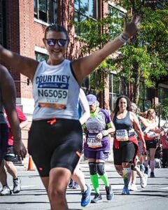 A smiling runner with arms raised crosses a city street during a race, wearing sunglasses and a bib labelled Courtney. Other runners follow behind, some wearing colourful athletic kit. Brick buildings and trees line the street.
