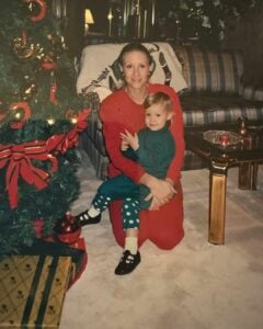 A woman in a red dress sits on the floor by a young child wearing green pyjamas, near a decorated Christmas tree and presents in a cosy living room. Both are smiling at the camera.