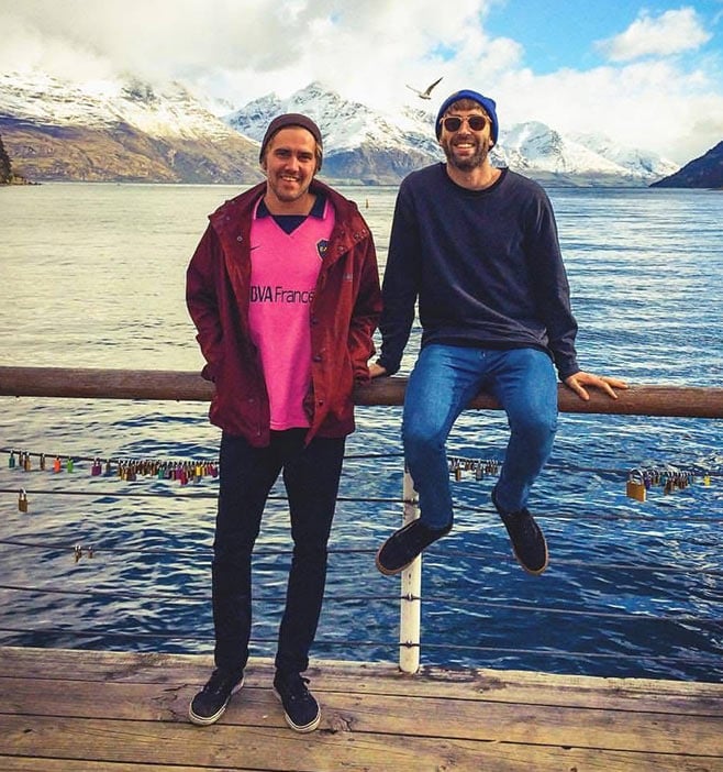 Two men in casual clothes and beanies pose on a wooden railing by a lake with snow-capped mountains in the background. One stands while the other sits, smiling. Padlocks are attached to the railing. A bird flies overhead.