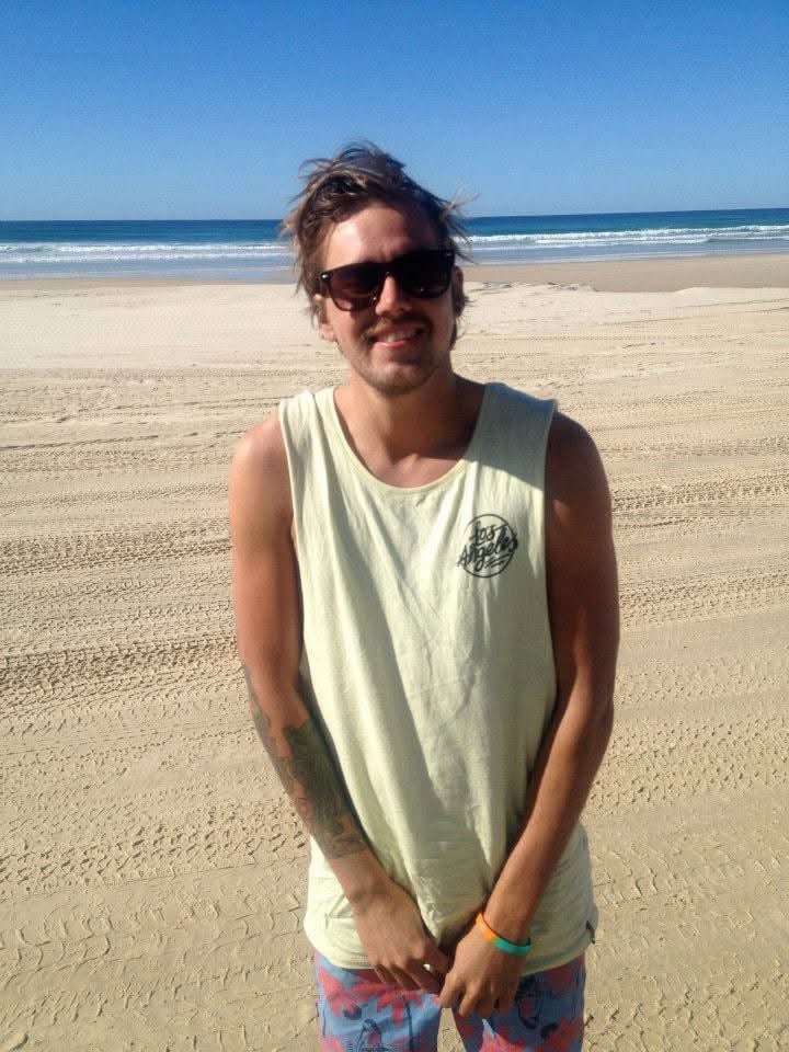 A young man wearing sunglasses, a sleeveless tan shirt, and patterned shorts stands on a sandy beach with the sea and blue sky in the background. He is smiling and has tousled hair.