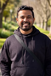 Abishek Anand: A man with short dark hair and a beard smiles outdoors. He is wearing a black hoodie and a crossbody strap, with a background of trees and sunlight.