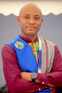 Kouamé Ambroise Kintossou: A man with a shaven head stands with arms crossed, wearing a maroon shirt and a colourful sash featuring blue, green, yellow, and striped patterns, and a black wristwatch. The background is blurred and light-coloured.