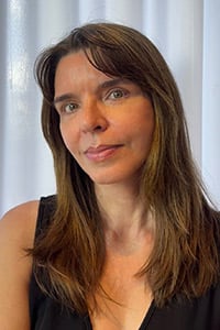 Ana Paula Drummond Lage: A woman with long brown hair and green eyes, wearing a sleeveless black top, stands in front of white curtains and looks at the camera with a relaxed expression.