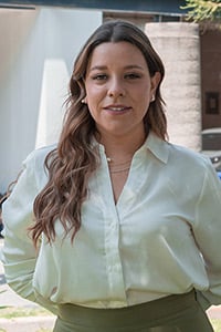Liliana Gómez Flores Ramos: A woman with long, wavy brown hair stands outdoors, wearing a light-coloured blouse and a necklace. She is looking at the camera with a neutral expression, and there are blurred buildings and greenery in the background.