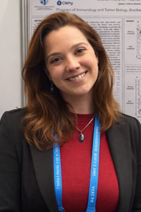 Jessica Magalhães Cotias Ristow Branco: A woman with shoulder-length brown hair, wearing a black blazer, red top, blue lanyard, and a necklace, smiles at the camera in front of a scientific poster display.