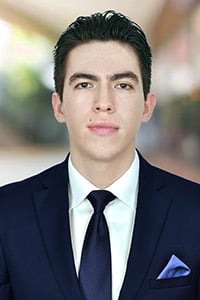 Jaime Ivan Castillo Silva: A young man in a navy blue suit, white shirt, and dark tie stands in front of a blurred indoor background, looking directly at the camera with a neutral expression.
