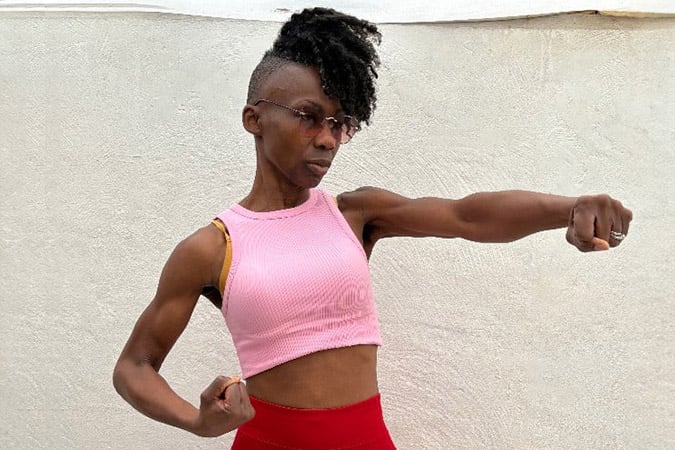 A person with a short curly hairstyle and glasses, wearing a pink crop top and red trousers, poses in a strong boxing stance against a plain white background—channelling the determination of Kemi Williams' London Marathon cancer fundraising story.