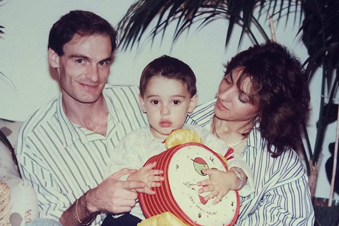 A man, a woman, and a young child sit together indoors. The child holds a red toy drum and looks at the camera, while the man smiles and the woman looks fondly at the child. There are plants in the background.