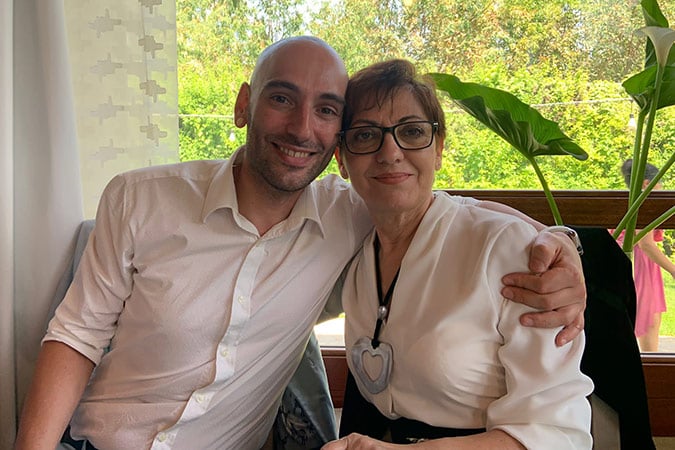 A man and an older woman, both wearing white shirts, sit closely together and smile at the camera. The man has his arm round the woman. They are indoors near a window with green plants visible outside.