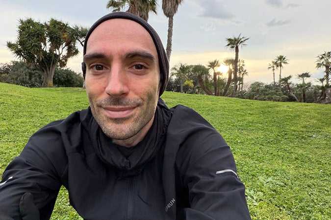 A person wearing a black jacket and beanie takes a selfie outdoors whilst sitting on green grass, with palm trees and a cloudy sky in the background at sunset.