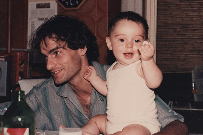 A man with dark hair and a striped shirt smiles and looks to his left while holding a baby in a white babygrow who is reaching forward and smiling. They are indoors, sitting at a table with bottles and cups.