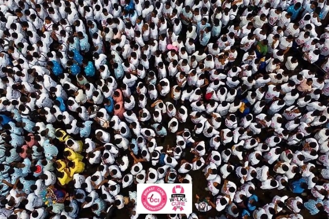A large crowd seen from above, many wearing white shirts, gathers for a breast cancer awareness walk. Several pink-themed logos about breast cancer in sub-Saharan Africa are visible at the bottom of the image.