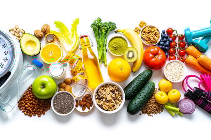 An assortment of healthy foods including fruit, vegetables, nuts, beans, grains, and seeds, arranged alongside a water bottle, measuring tape, scales, and colourful dumbbells and skipping rope, all on a white background.