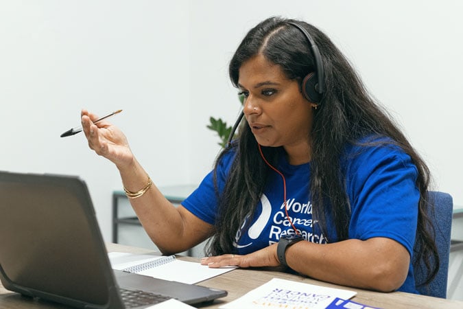 A woman wearing a headset and a blue World Cancer Research shirt sits at a desk, looking at a laptop, holding a pen, and taking notes in a notebook whilst preparing for cancer prevention workshops for health professionals.