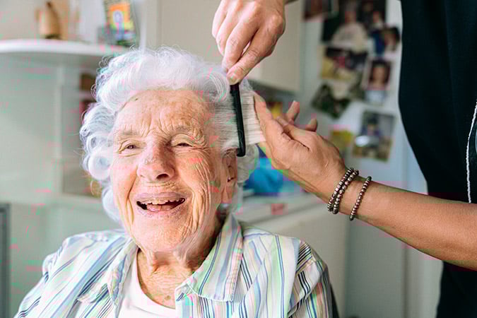 An elderly woman with white hair smiles joyfully as another person gently brushes her hair. The setting appears to be a cosy home with family photos on the wall in the background.