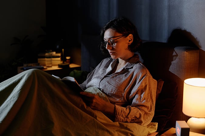 A woman sits up in bed at night reading a book. It's nighttime and a bedside lamp is on.