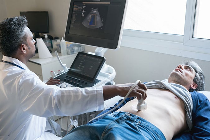 A doctor performs an abdominal ultrasound on a male patient lying on an examination couch, while viewing the scan results on a monitor.