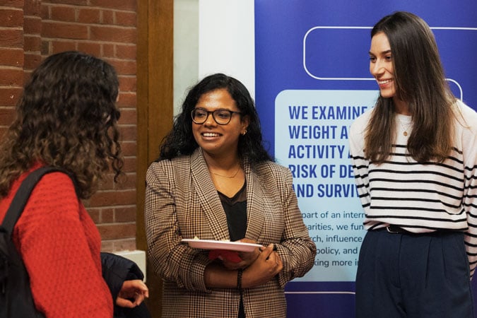 Three women are standing and talking indoors, one holding a notebook, another in a striped jumper, and the third in a red jumper. They appear to be at an event with a blue informational poster about cancer prevention resources for health professionals behind them.
